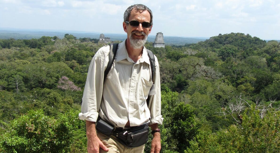 En mand iført solbriller og vandreudstyr smiler til kameraet. Han står foran en frodig, grøn skov med gamle stenstrukturer synlige i baggrunden under en overskyet himmel.