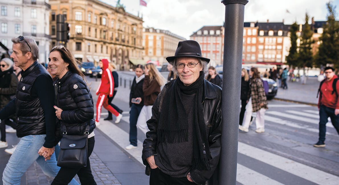 En mand i hat og halstørklæde står på en bygade i nærheden af et krydstogt. Omkring ham går fodgængere, nogle i vintertøj. Baggrunden viser historiske bygninger under en delvist overskyet himmel.
