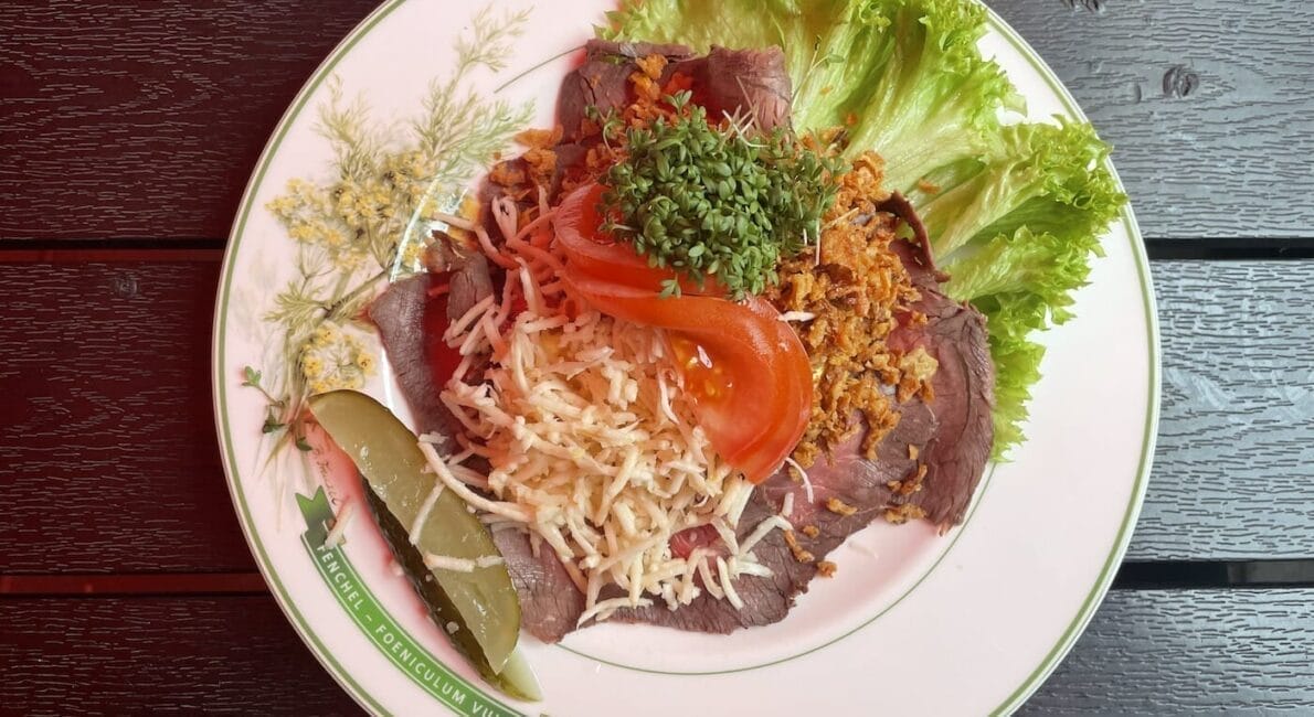 En tallerken med oksekød i tynde skiver toppet med revet ost, stegte løg, tomatskiver og en lille bunke grønt. Pyntet med salat og en agurk skive, serveret på en dekorativ tallerken.