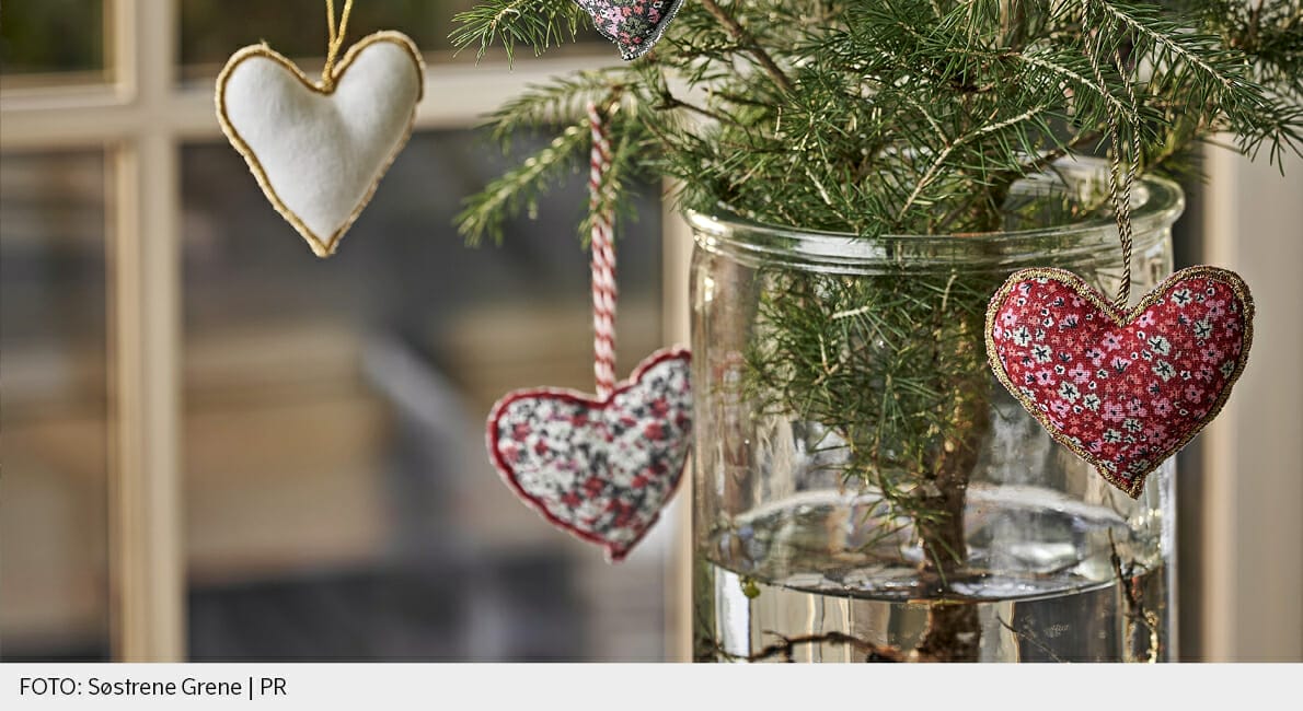 Nærbillede af et lille fyrretræ i en glasvase fyldt med vand. Tre hjerteformede ornamenter hænger fra grenene, hver med forskellige stoffer: hvid, blomstret og rød med print. Et vindue er sløret i baggrunden.
