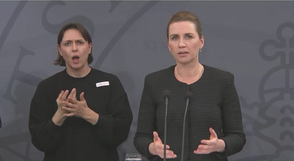 En kvinde i mørkt tøj taler ved et podium med mikrofoner. Ved siden af hende bruger en anden kvinde i lignende påklædning tegnsprog. Baggrunden har et subtilt, abstrakt design.