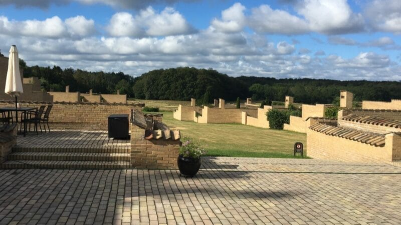 Gårdhave med stengulv og murstensvægge overset af et landskab med grønne marker og træer. Der er udendørs borde og stole til venstre, skyer på den blå himmel og en potteplante i forgrunden.