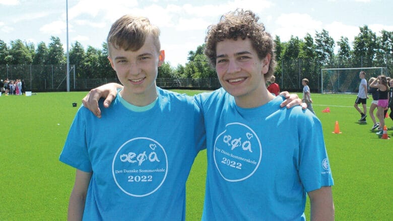 To teenagedrenge i blå t-shirts med tekst, smilende, mens de poserer på en sportsplads. Der er en gruppe, der spiller i baggrunden under en delvist overskyet himmel.