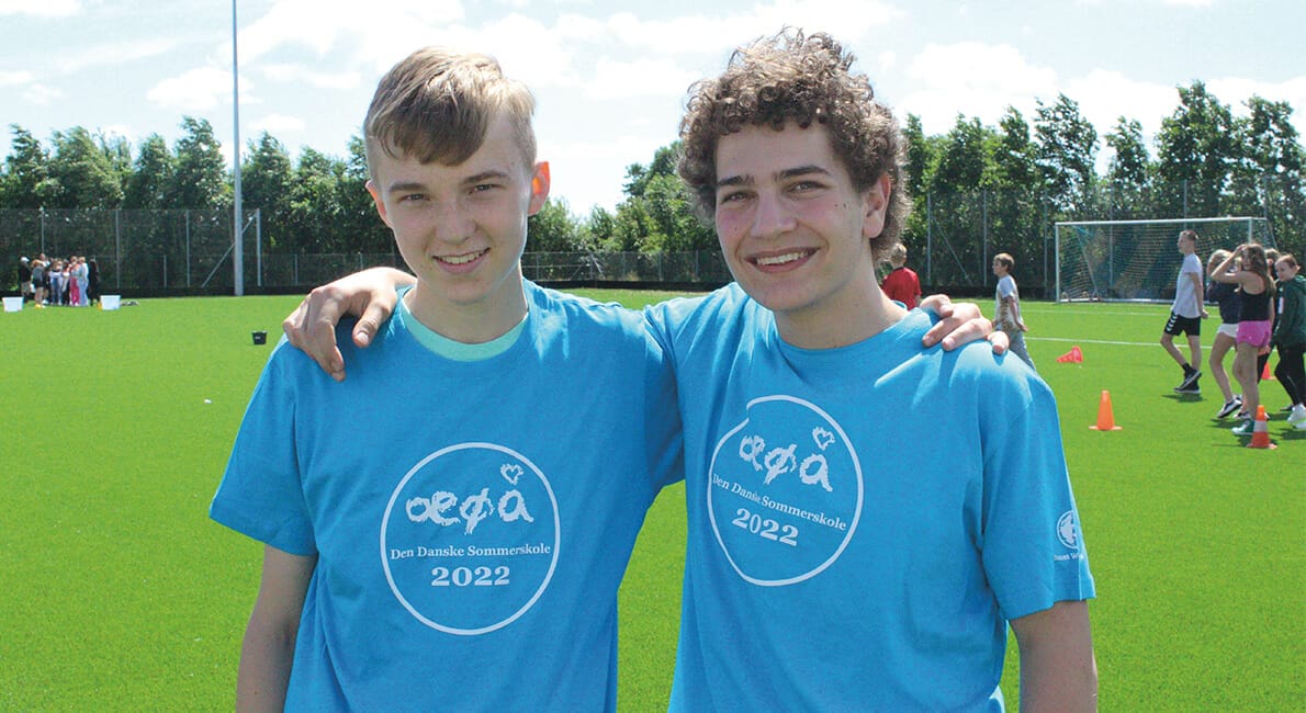 To teenagedrenge i blå t-shirts med tekst, smilende, mens de poserer på en sportsplads. Der er en gruppe, der spiller i baggrunden under en delvist overskyet himmel.