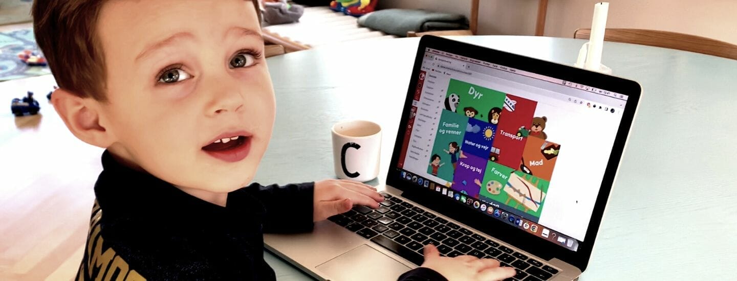A young child is sitting at a table using a laptop. The computer screen displays colorful graphics with cartoon characters. A mug with the letter C is beside the laptop. The background is a softly lit room with toys and a couch.