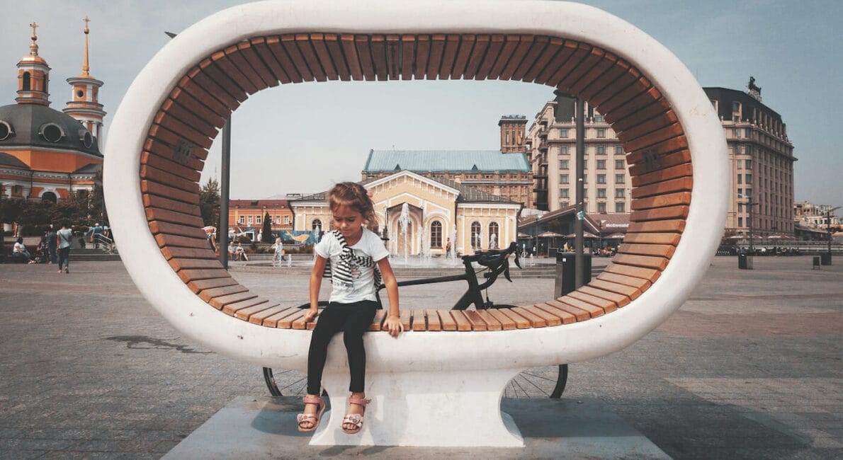 A child sits on a large, uniquely shaped circular wooden bench in a city square in Ukraine. The cityscape features historical buildings, including a church with golden domes. A bicycle is parked behind the bench as the sky remains overcast.
