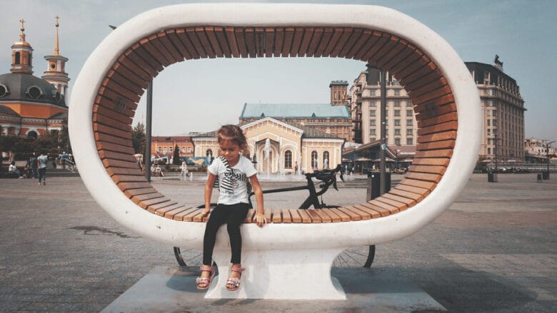 A child sits on a large, uniquely shaped circular wooden bench in a city square in Ukraine. The cityscape features historical buildings, including a church with golden domes. A bicycle is parked behind the bench as the sky remains overcast.