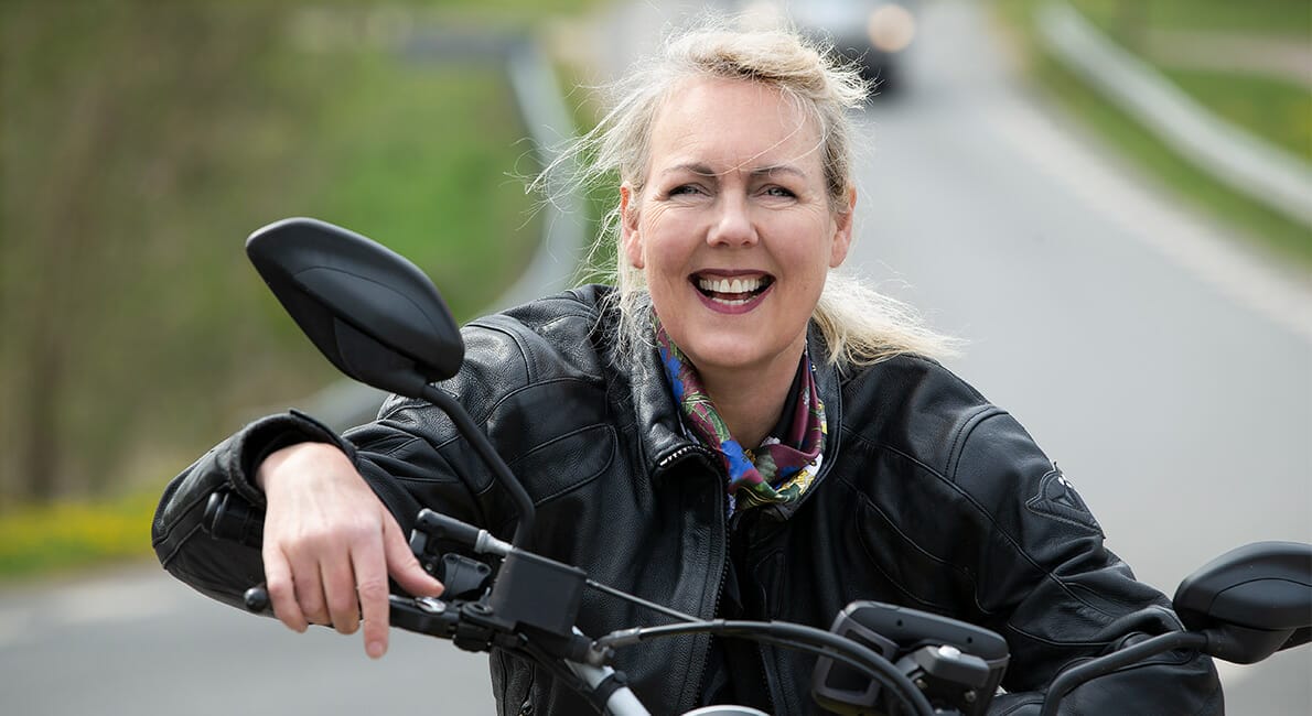 Smilende kvinde iført en sort læderjakke og farverigt tørklæde kører på motorcykel ned ad en landevej. Baggrunden er sløret og viser grønne områder og et andet køretøj i det fjerne.