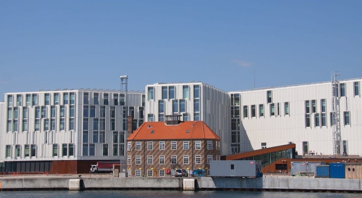 A modern waterfront building complex with large glass windows, incorporating a smaller historic building with a red-tiled roof. The scene is set against a clear blue sky and calm water.