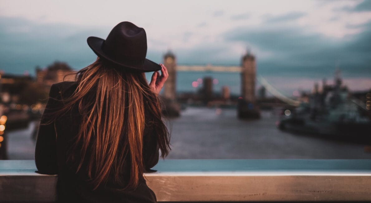 En person i mørk frakke og hat står på en bro og stirrer på Tower Bridge i London i en overskyet tusmørke. Floden flyder nedenunder, byens lys begynder at oplyse scenen og fanger det reflekterende øjeblik af en studerende i Storbritannien.