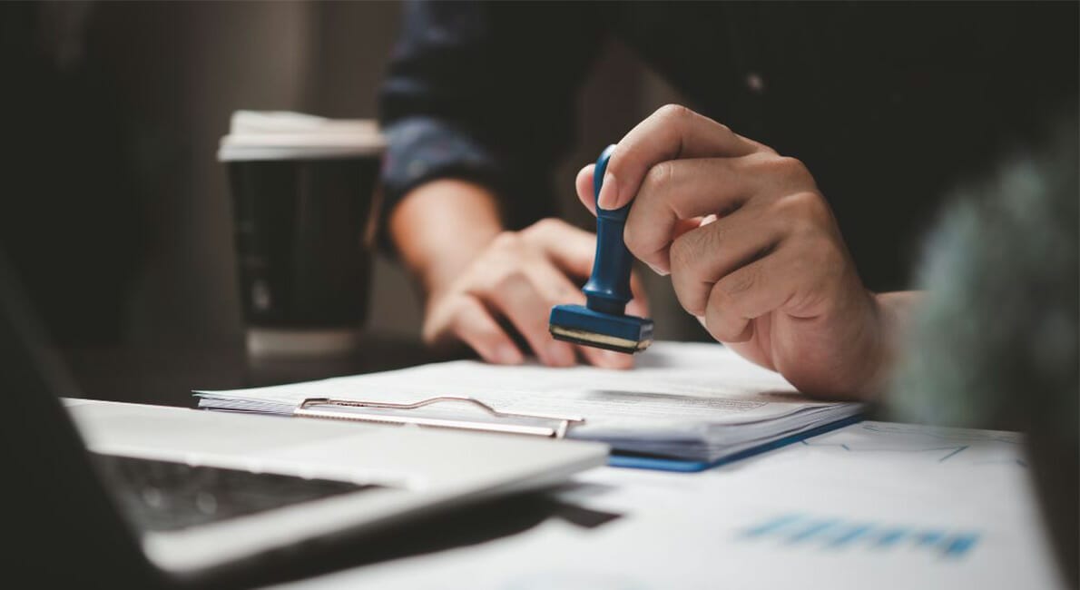 En person holder et gummistempel over et dokument på et skrivebord. En udklipsholder med papirer, en bærbar computer og en kaffekop er i nærheden. Indstillingen foreslår et kontormiljø med fokus på administrative eller godkendelsesaktiviteter.