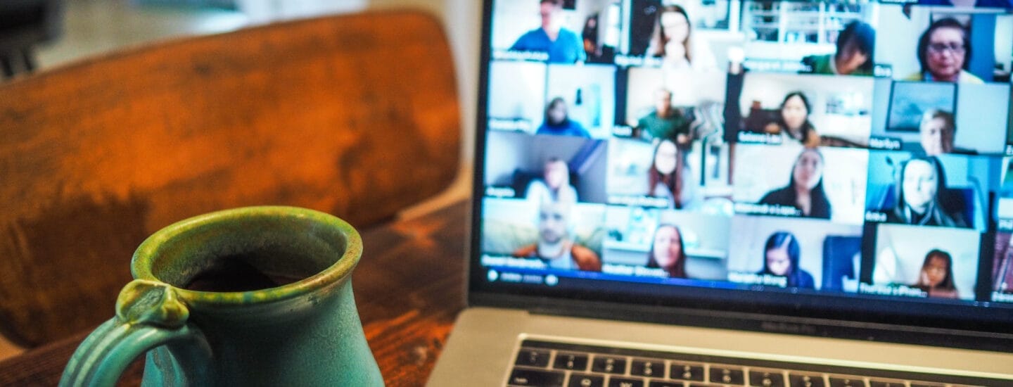 En bærbar computer sidder på et træbord og viser et gitter af deltagere i et videoopkald. Ved siden af den bærbare computer er der et keramisk krus med et håndtag. Indstillingen foreslår et hjem eller et hyggeligt arbejdsmiljø.