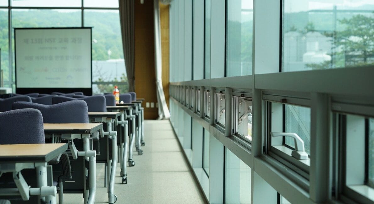 Et moderne mødelokale med rækker af blå stole og foldbare borde ud mod et projektorlærred. Store vinduespartier til højre giver udsigt til grønne områder udenfor. Naturligt lys fylder rummet.