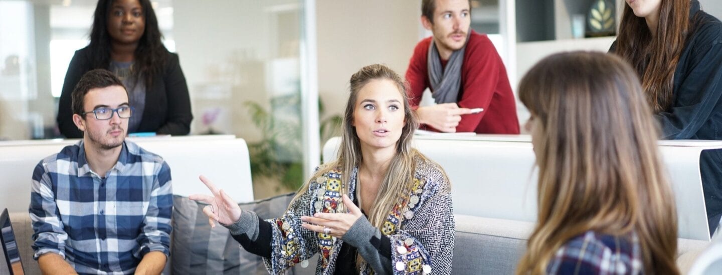 En gruppe på seks personer har en diskussion i et moderne kontormiljø, hvor de udforsker muligheder for virksomhedspartnerskaber. En kvinde i en mønstret jakke taler, mens de andre lytter opmærksomt, omgivet af bærbare computere og notesblokke, der understreger deres samarbejdssession.