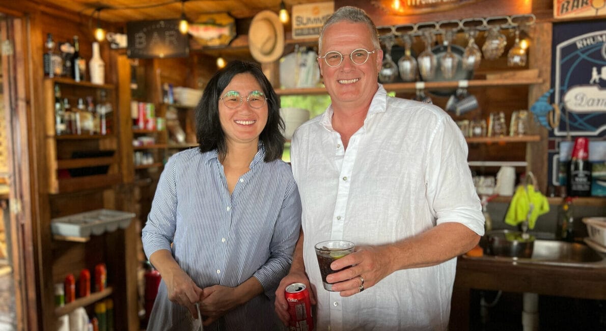 En kvinde og en mand smiler bag en bardisk i træ. Kvinden er iført briller og en stribet skjorte. Manden er iført briller og hvid skjorte og holder en drink og en dåse. Baren har forskellige flasker og glas i baggrunden.