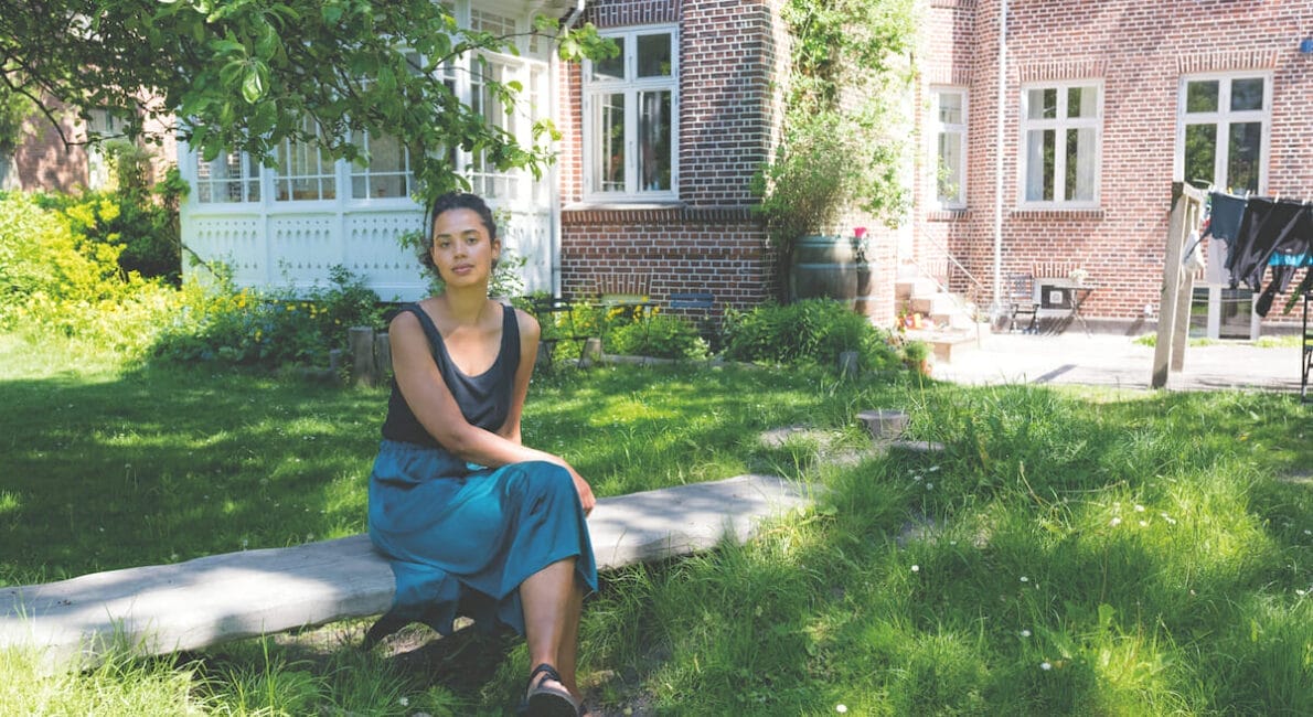 En kvinde sidder på en træbjælke i en have ved siden af et rødt murstenshus med hvidindrammede vinduer. Hun bærer en mørk top og en lang blå nederdel, med sandaler, midt i frodige grønne områder og sollys, der filtrerer gennem træerne.